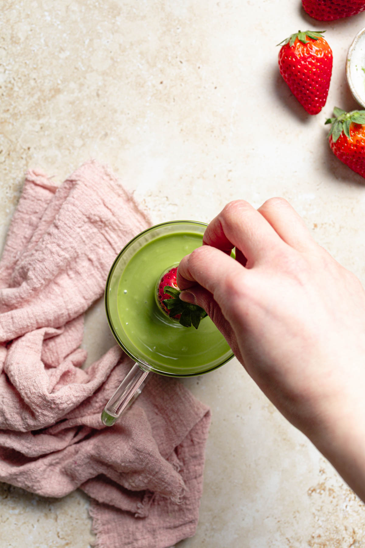 A hand dipping a strawberry into melted matcha chocolate